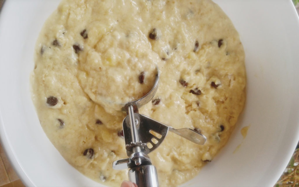 An ice cream scoop scoops out muffin batter from a white bowl.