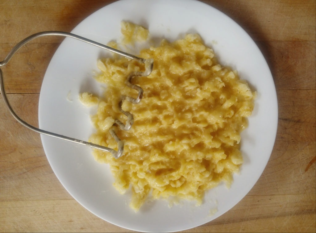 A white plate of mashed bananas. A potato masher sits on the plate.