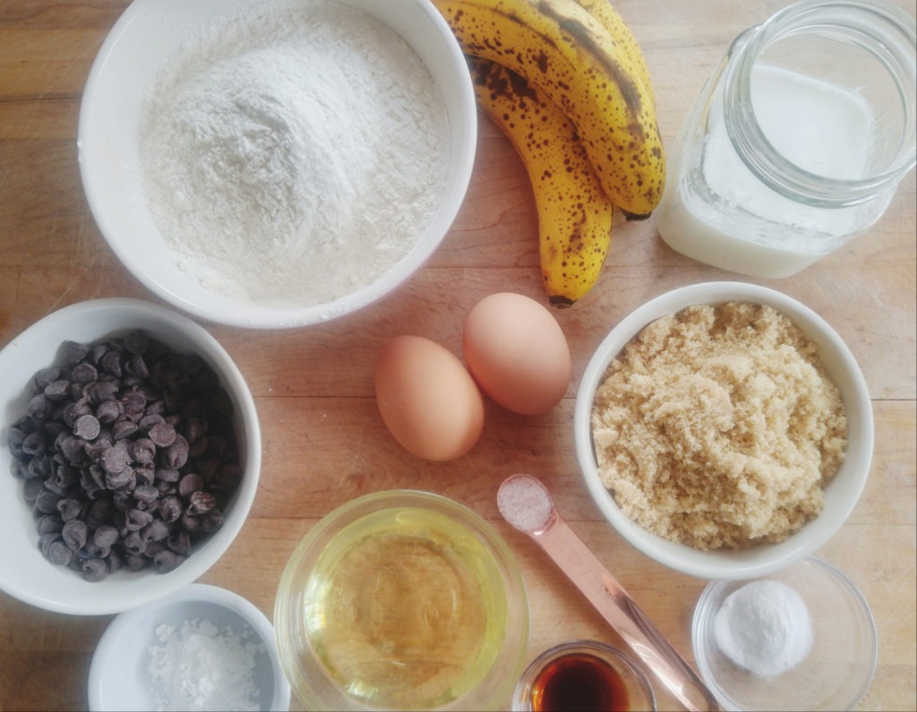 A cutting board displaying muffin ingredients, including bananas, eggs, brown sugar, chocolate chips, salt, flower, and milk in a jar.