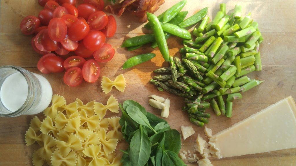 A cutting board is filled with piles of chopped asparagus, fresh parmesan cheese, basil, raw bowtie pasta, bacon, halved cherry tomatoes, and a mason jar of cream.