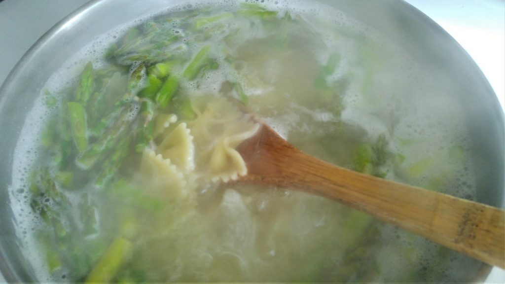 A pot full of boiling water and chopped asparagus and bowtie pasta. It is being stirred with a wooden spoon.