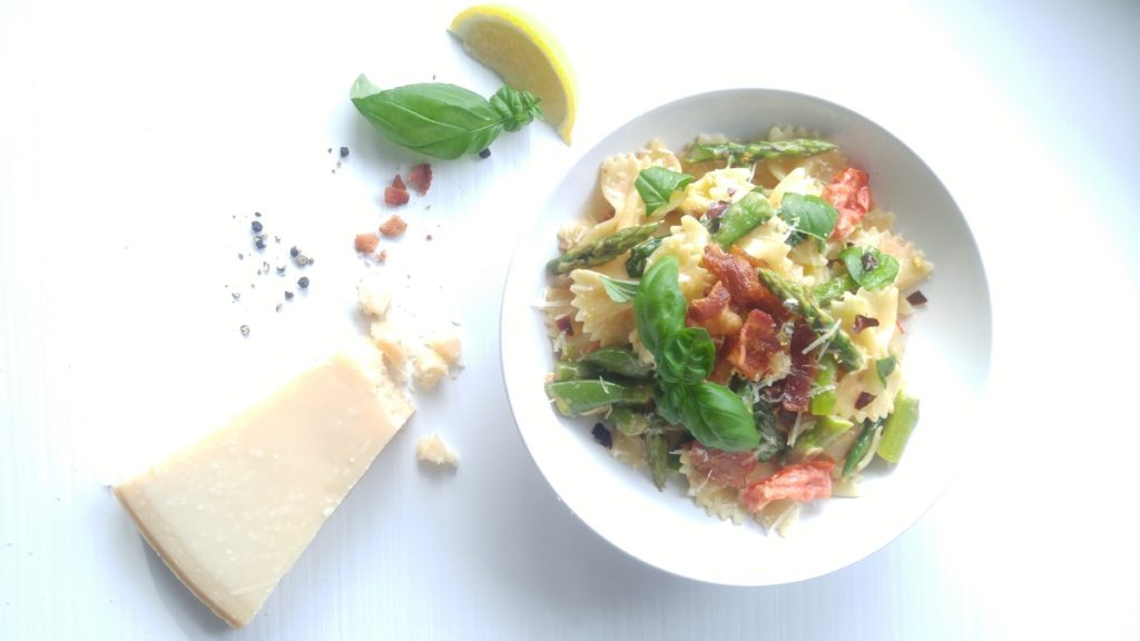 White serving bowl is filled with a blend of bowtie pasta, asparagus, bacon, and basil. A block of parmesan cheese sits on the table beside it.