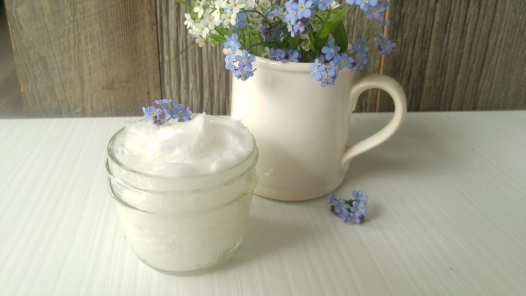 A small jar of moisturizer sits on a table with a small white vase of light blue and white flowers.