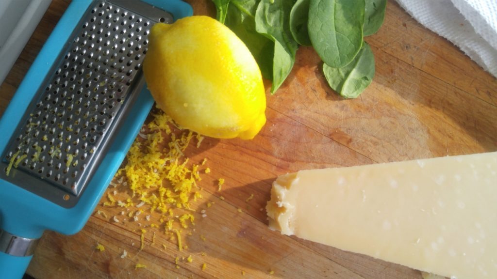 A close up picture of a cutting board with a zested lime, basil, and a block of parmesan cheese.