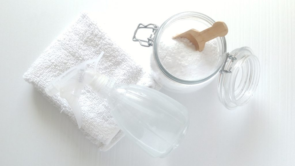 A clear jar of white powder, a white cloth, ann a spray bottle sit on a white table