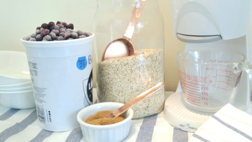 Frozen blueberries, raw oats, and coconut sugar on a table beside a hot water maker and some ceareal bowls and a measuring cup.