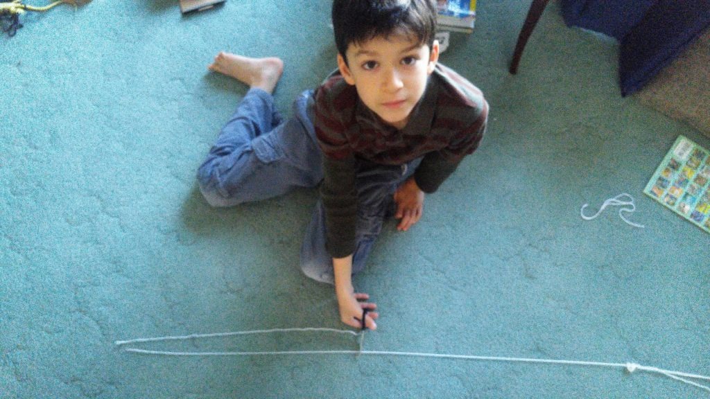 A young boy looks up at the camera, while demonstrating how to cut a long string of yarn in to smaller pieces.