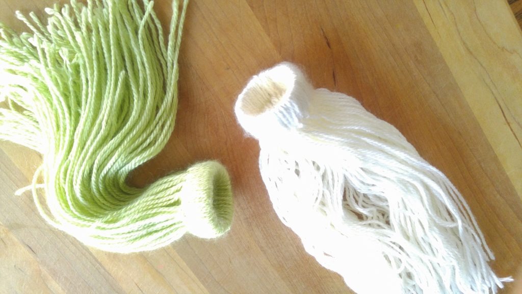 A white toque and green toque lie on the table. They are almost complete, but the strands of yarn are loose and need to be tied in to the shape of the toque.