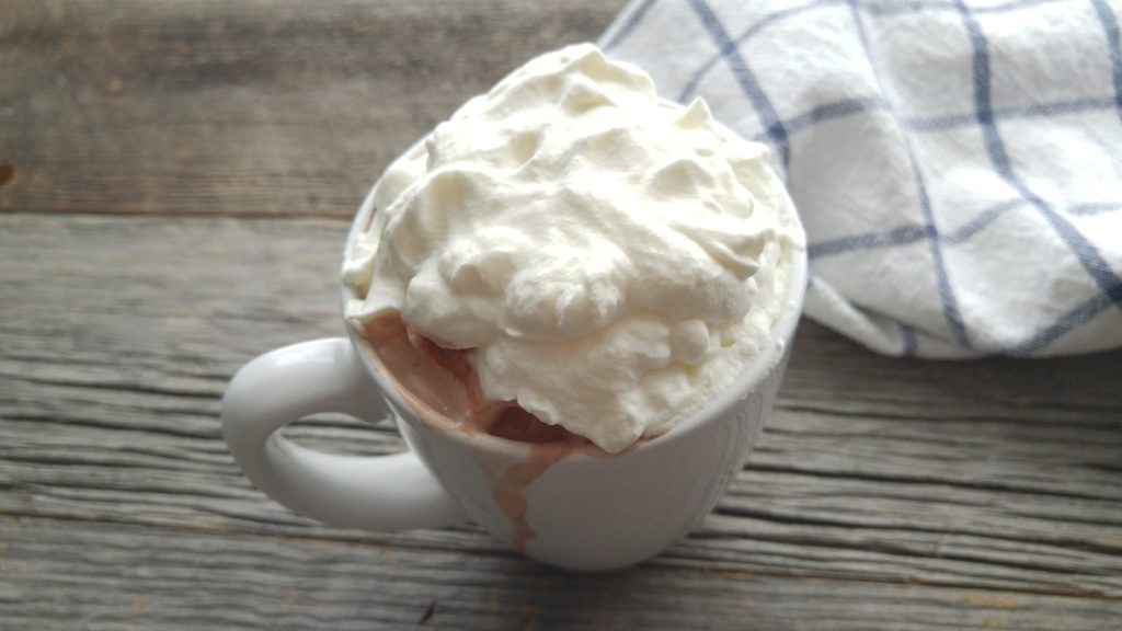 White mug of hot chocolate covered in whipped cream. Some hot chocolate is dripping down the outside of the mug.