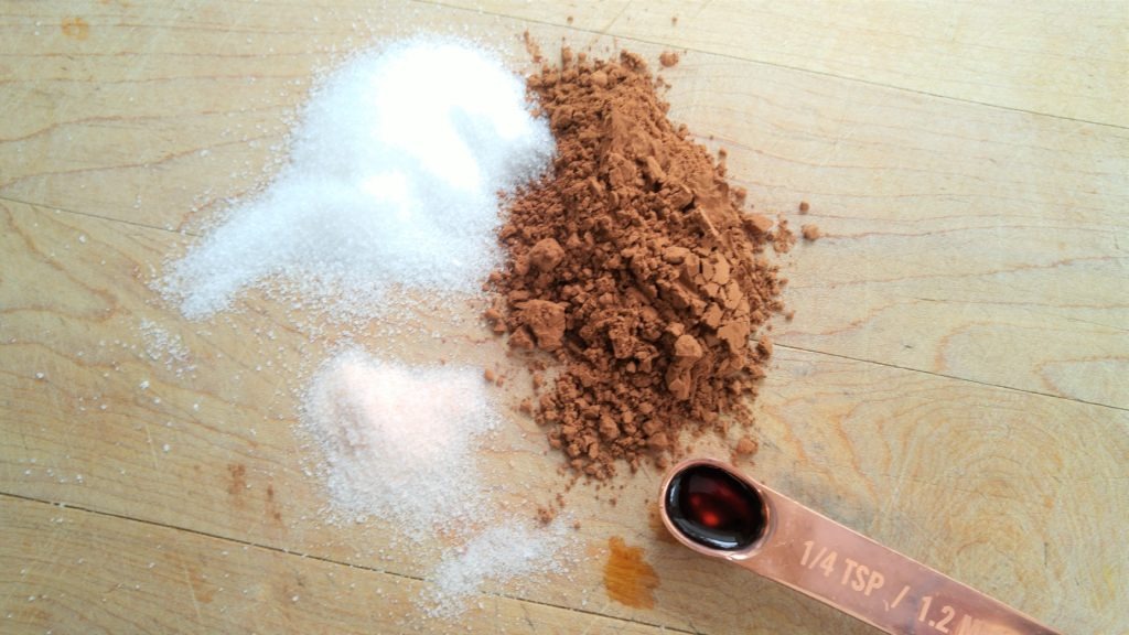 Small piles of cocoa, sugar, salt, and a measuring spoon of vanilla on a wooden cutting board.