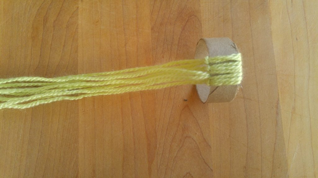 Multiple strands of green yarn are tied onto a round of toilet paper roll.
