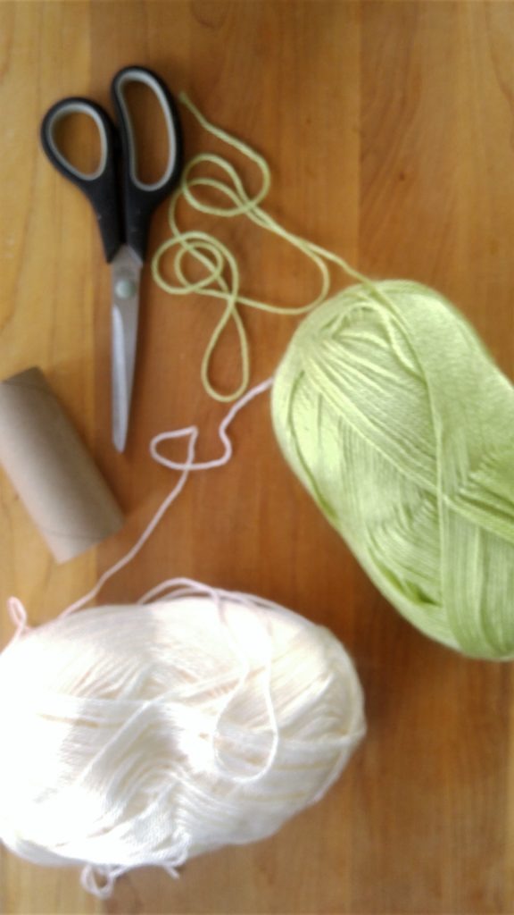 Balls of green and white yarn sit on a table with some scissors and a cardboard toilet paper roll.