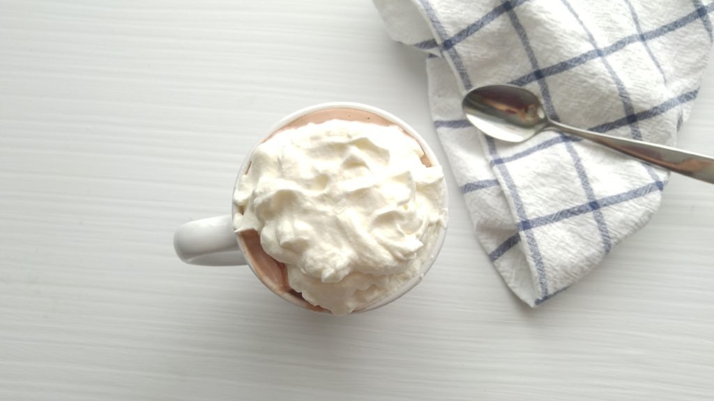 White cup of hot chocolate sits on a white table beside a teaspoon and a tea towel.