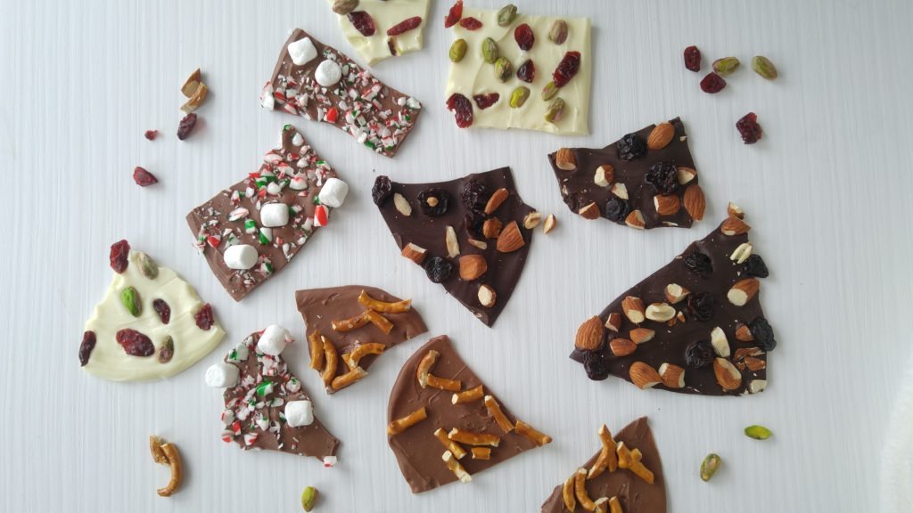 A white table with pieces of chocolate bark all over it. Some bark has almonds on it, others have pretzels, and others have cranberry and pistachios. Another has crushed candy cane pieces and miniature marshmallows.