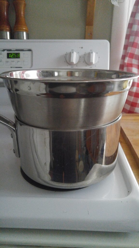 Close up of a double boiler. It is a saucepan sitting on a stove, with a stainless steel bowl resting on top of it.