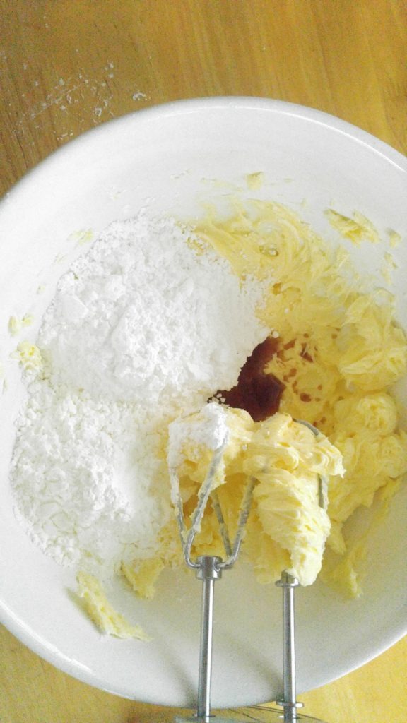 Close up of whipped butter, icing sugar and vanilla in a white bowl with beaters for mixing.
