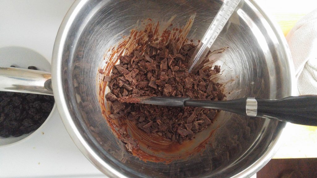 A stainless steel bowl has melting chopped chocolate in it, as well as a candy thermometer and a spatula which is stirring the chocolate.
