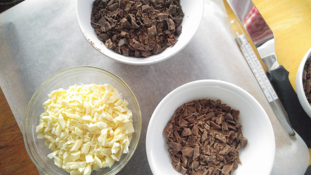 Three bowls of chopped chocolate, dark, white, and milk, sit on a parchment-lined cutting board.