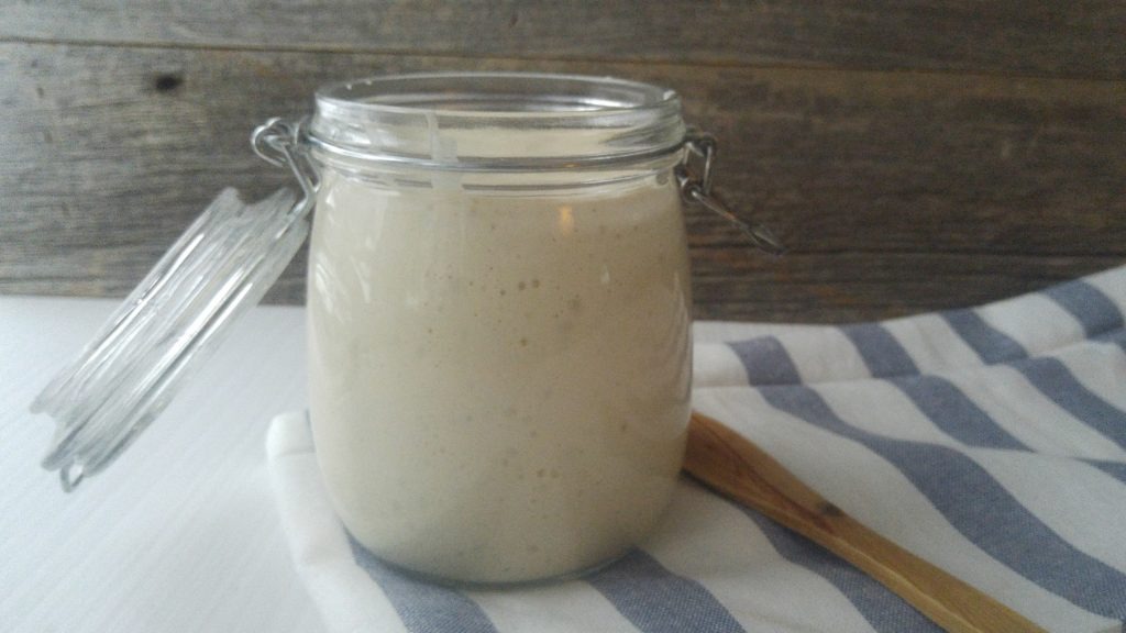 open glass jar full of fermenting sourdough starter. Placed beside a dish towel and wooden spoon on a white table.