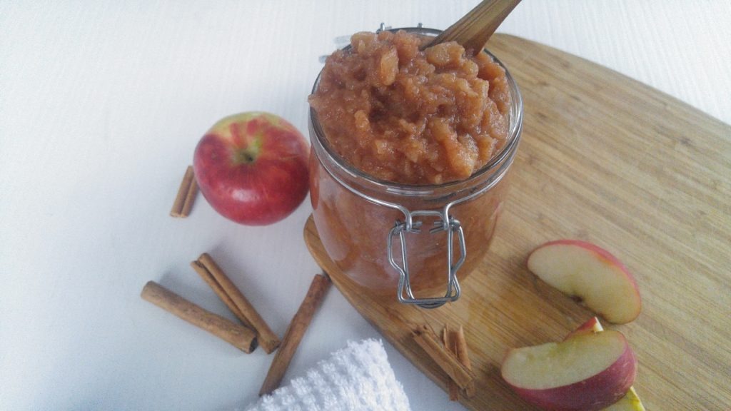 A jar filled with cinnamon applesauce sits on a cutting board surrounded by slices of apples and cinnamon sticks
