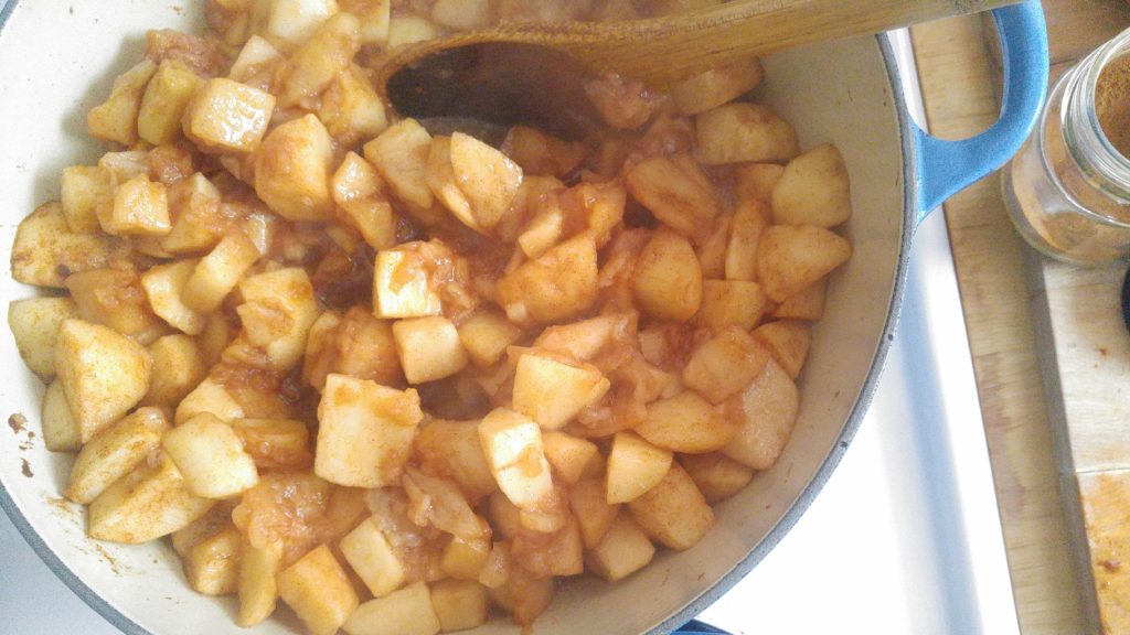 a wooden spoon is being used to stir apples in a sauce pan that are cooking down and looking softened.