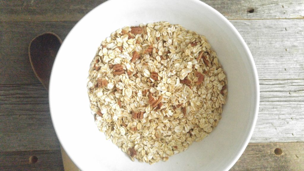 dry ingredients for granola in a white bowl including oats and pecans. It is set on a wooden table beside a wooden spoon.