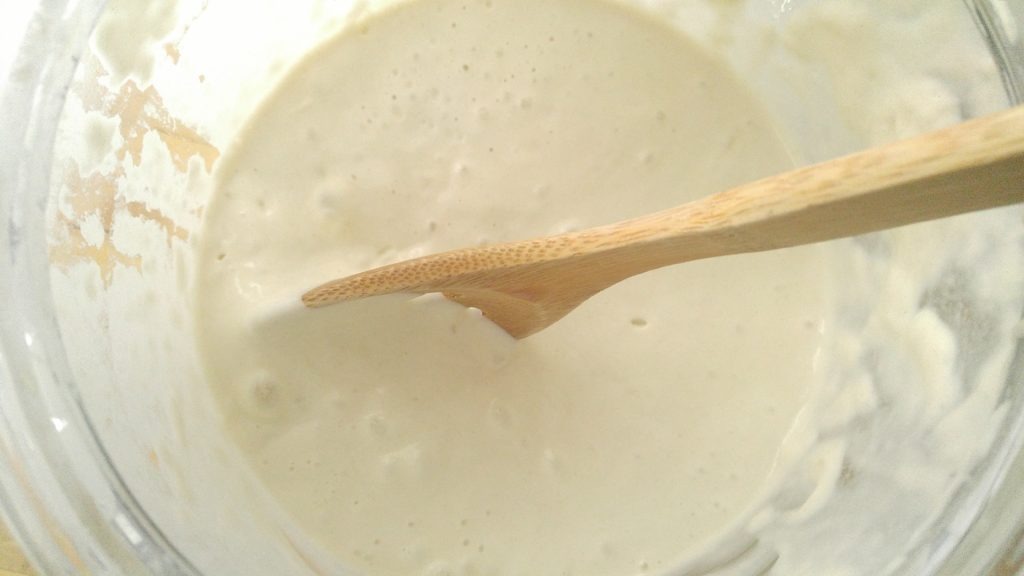 close up of making sourdoug starter with a wooden spoon stirring the liquid starter in a large glass container