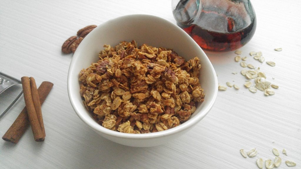 white cereal bowl of maple pecan granola on a white table beside maple syrup, cinnamon sticks, some pecans, and oats