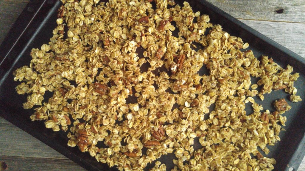 uncooked granola spread out thinly on a dark baking sheet, placed on a wooden table