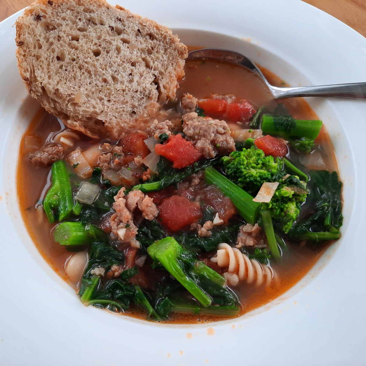 close up of hearty bowl of sausage and white bean soup full of rapini and tomato with a piece of multigrain bread beside it