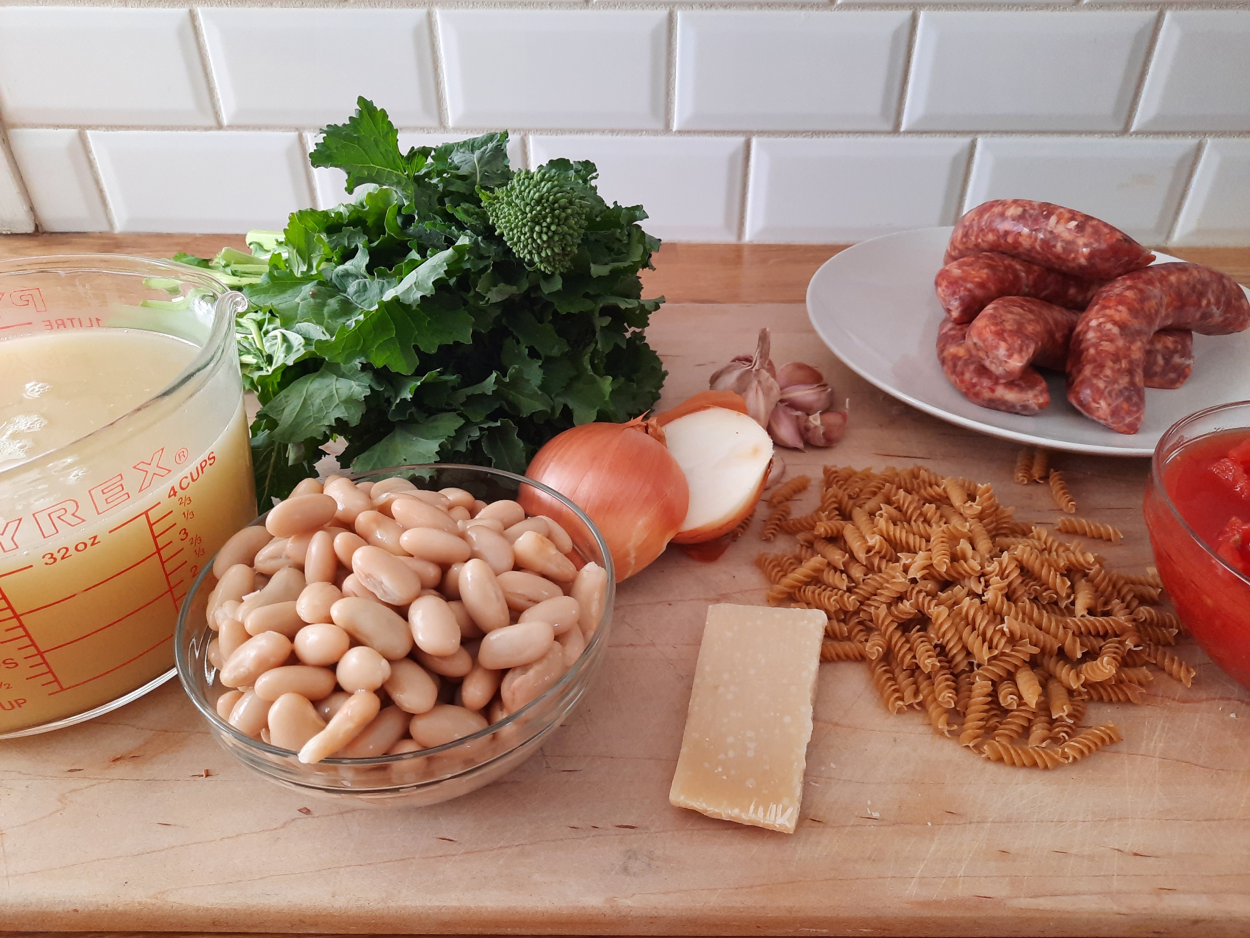 ingredients for soup on a cutting board, including beans, rapini, sausage, pasta, broth