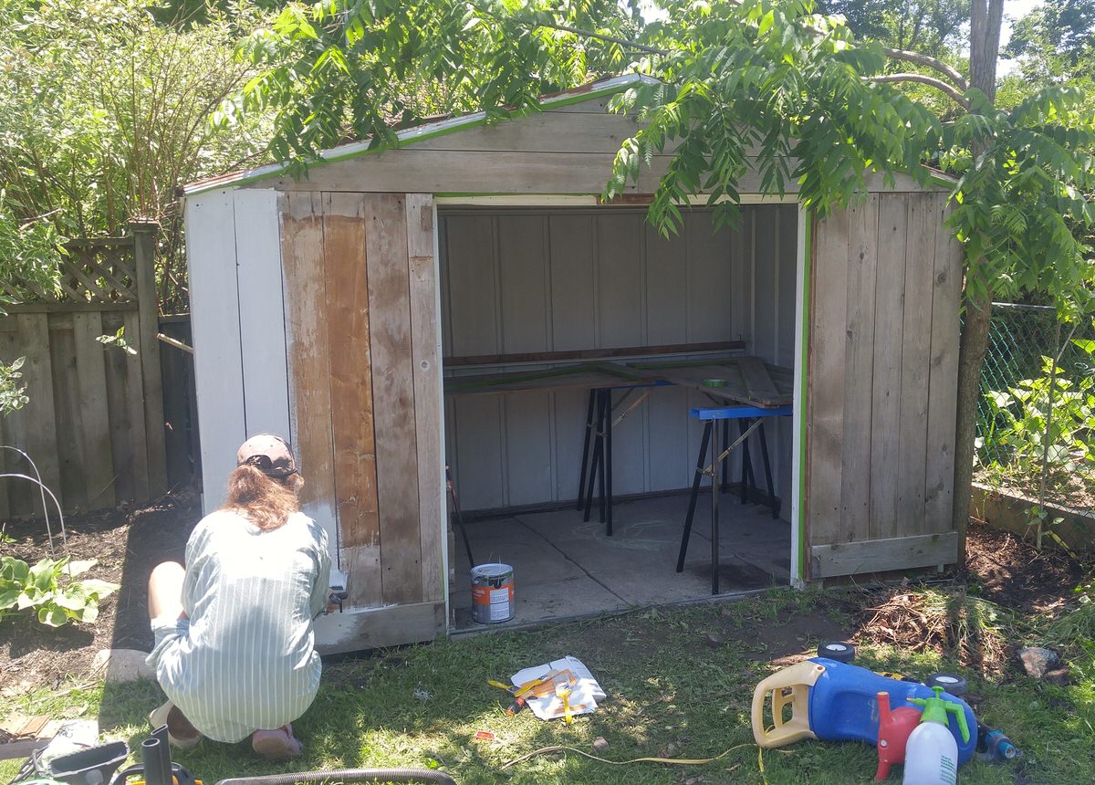 a woman bends down to paint a wooden shed wall