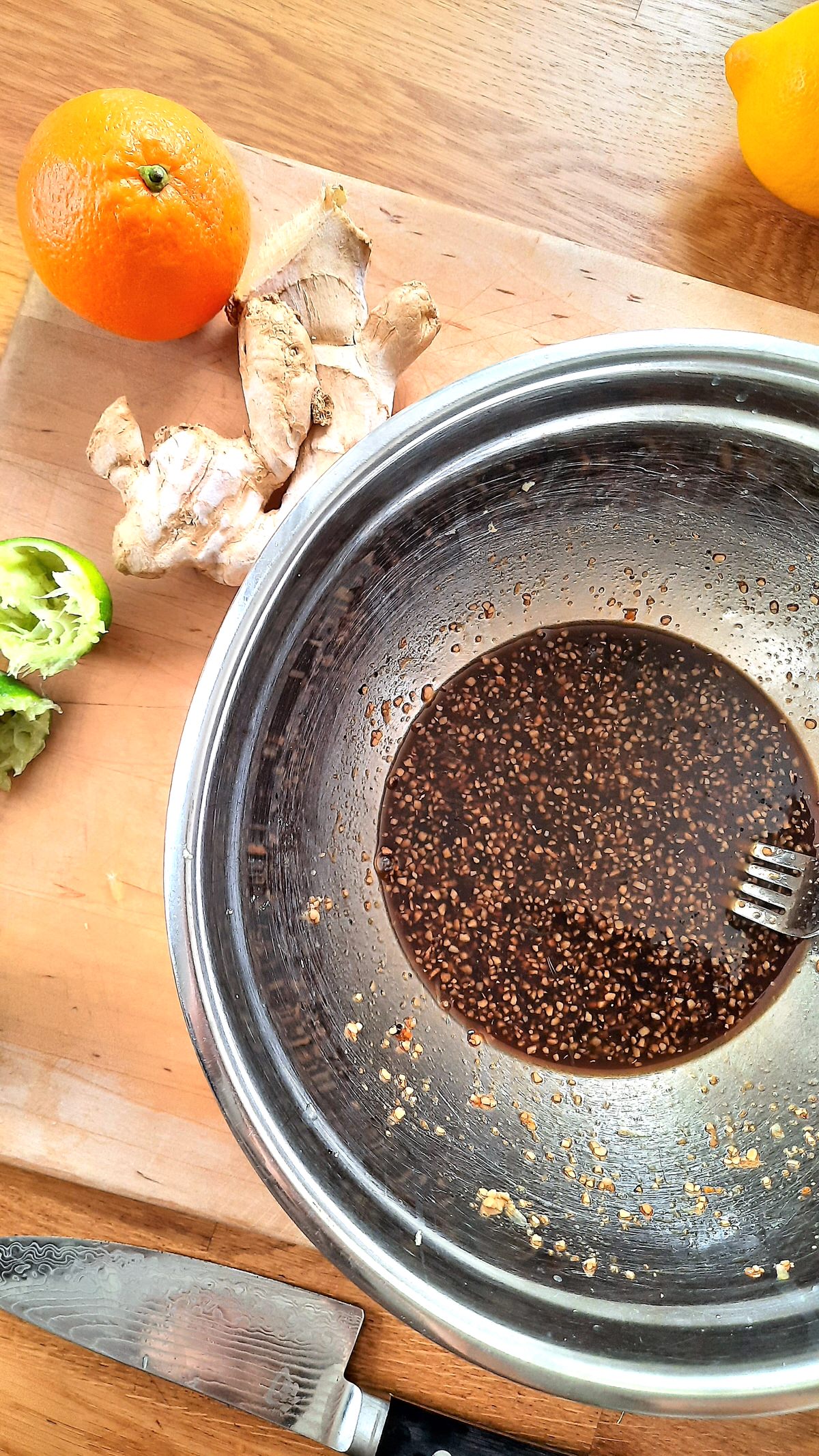 a bowl filled with rich brown chicken drumstick marinade on a counter beside pieces of ginger and orange segments