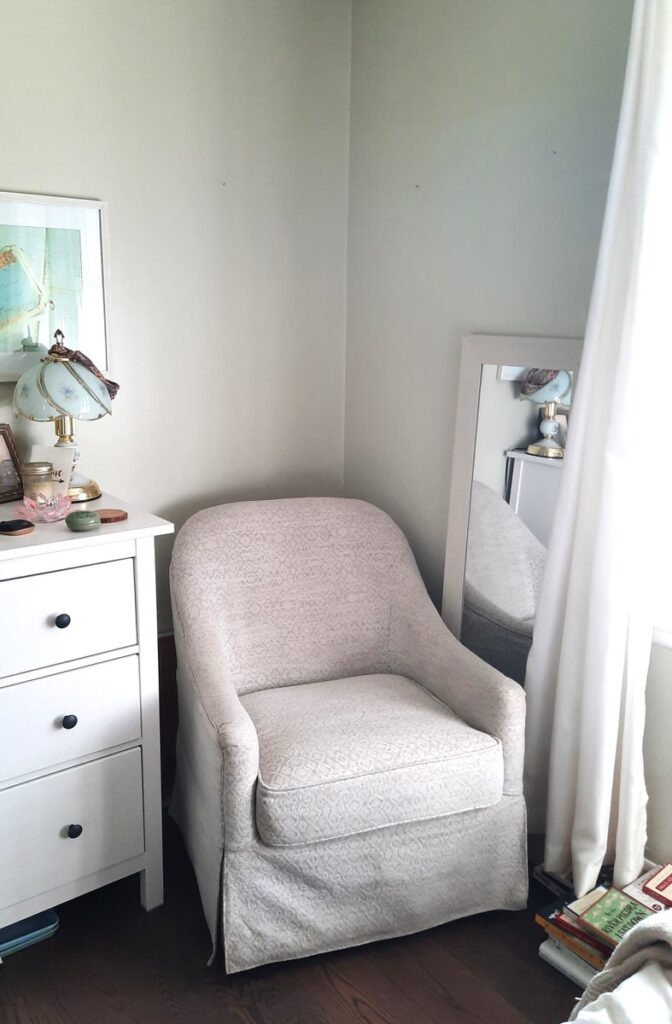 corner of a bedroom with a white dresser and white upholstered chair beside a window