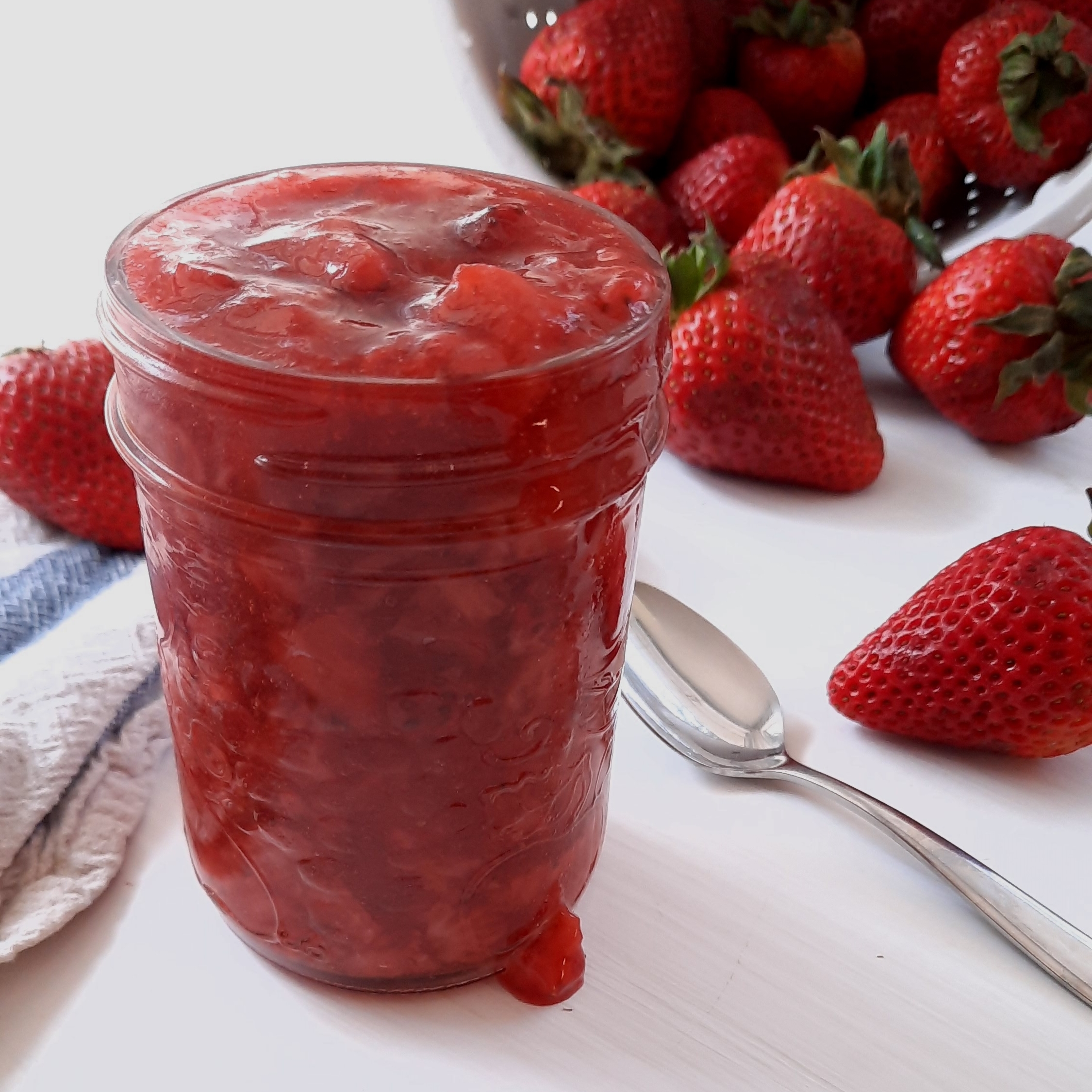 a jar filled with strawberry sauce overflows onto a white counter with whole strawberries surrounding it