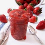 a jar filled with strawberry sauce sits on a white counter surrounded by fresh whole berries