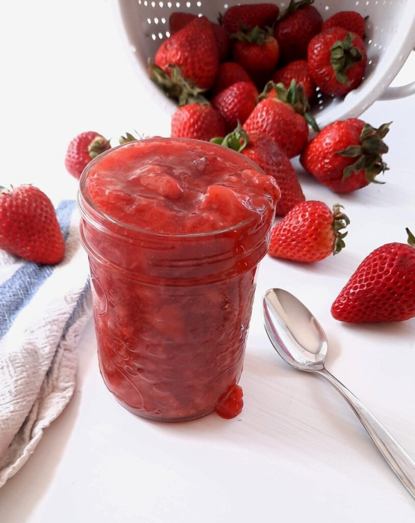 a jar filled with strawberry sauce sits on a white counter surrounded by fresh whole berries