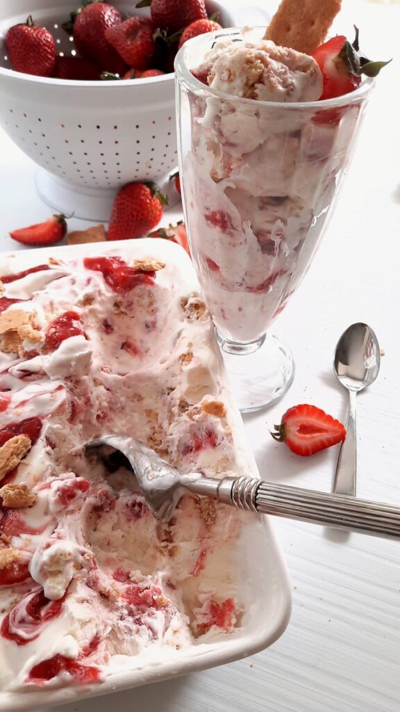 a loaf pan full of strawberry cheesecake ice cream sits beside a milkshake glass full of ice cream and a colander of fresh strawberries