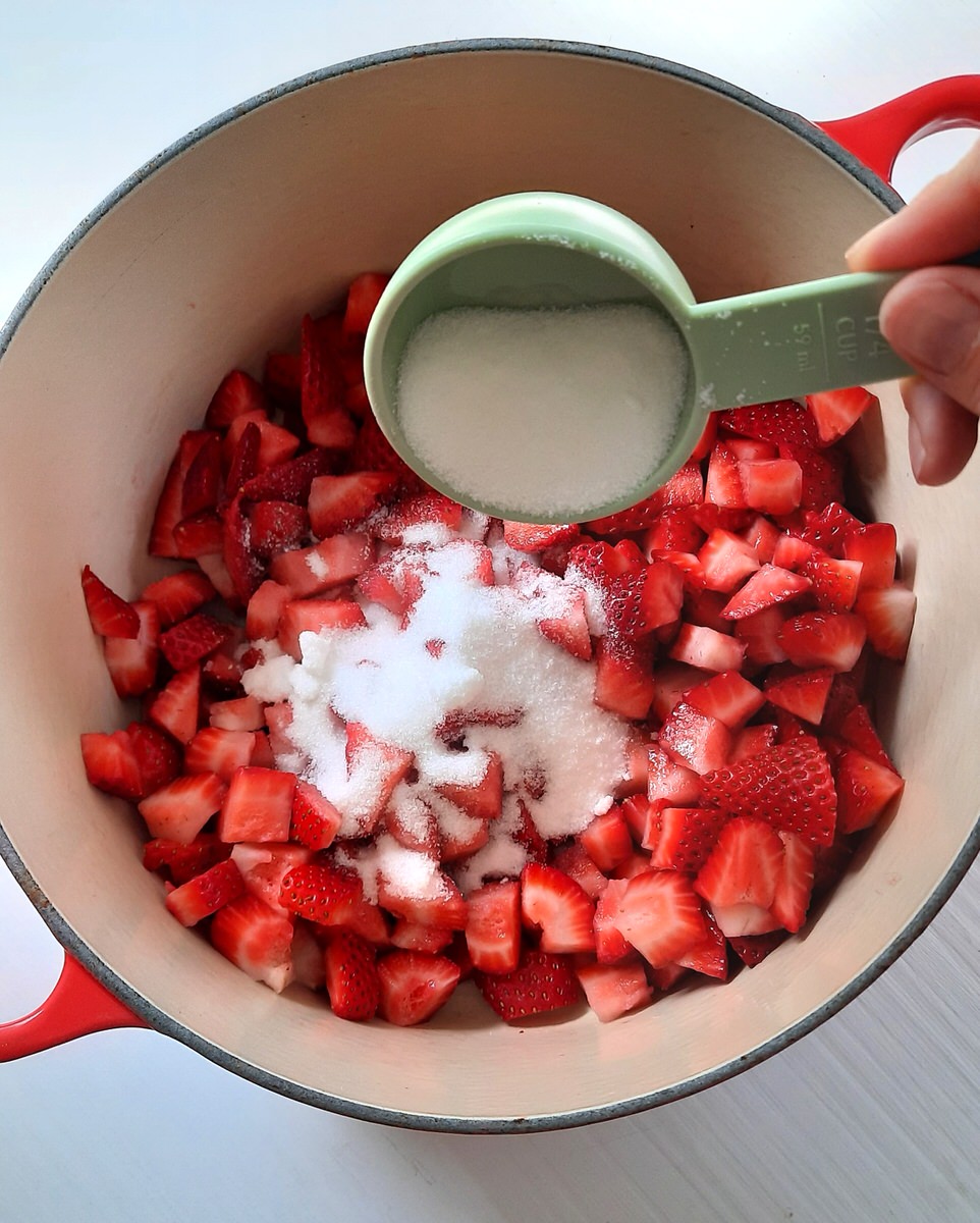 pouring sugar from a measuring cup into a pot of chopped strawberries