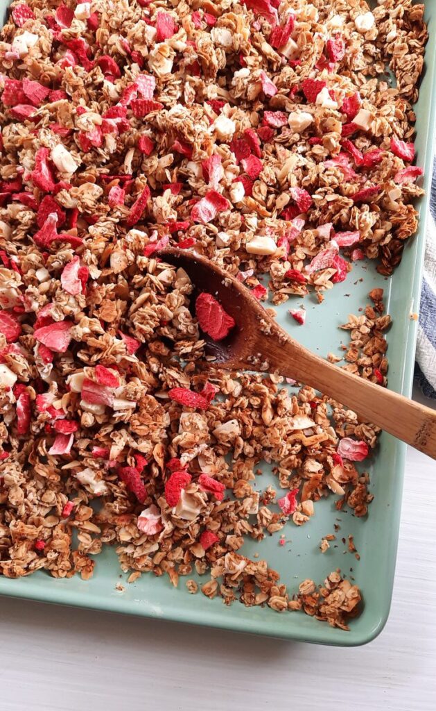 wooden spoon scooping strawberries & cream granola off of a baking sheet