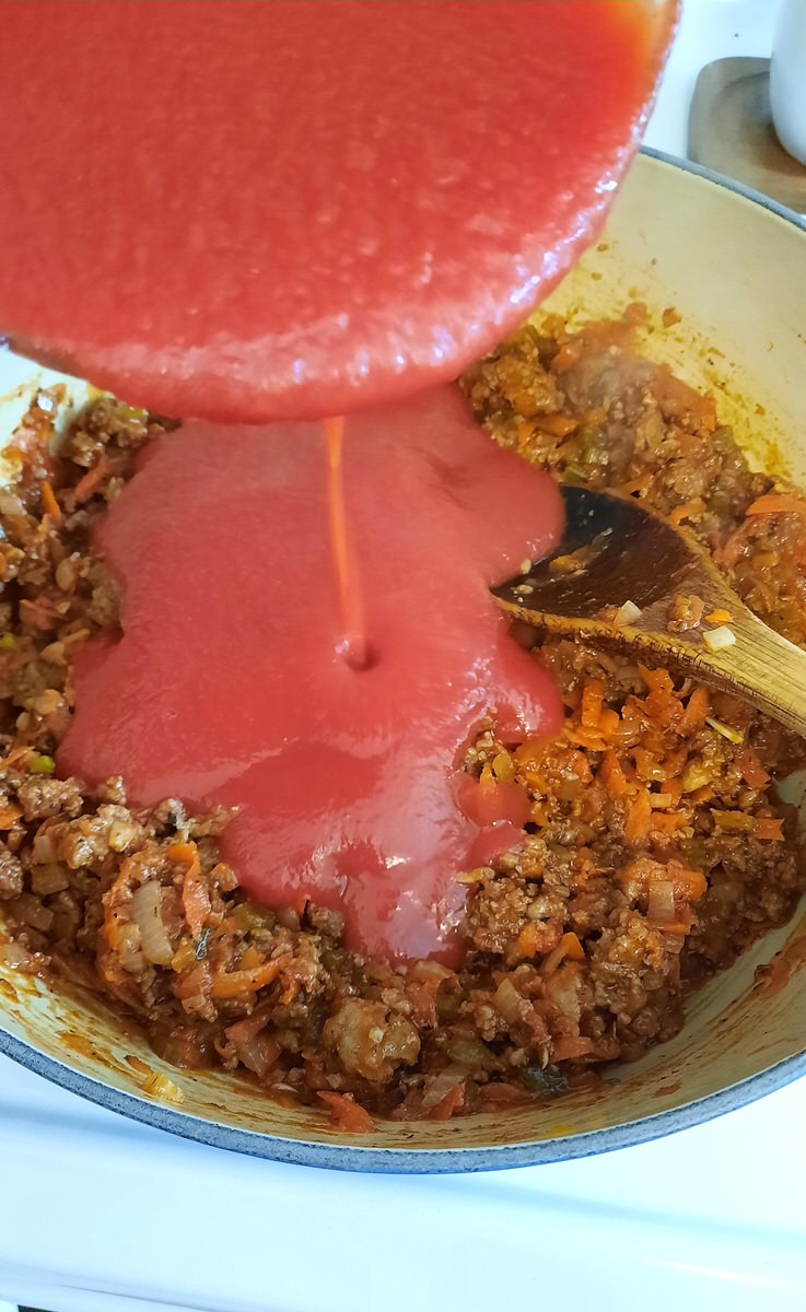 pouring tomato sauce into a pot of cooked ground meat and veggies