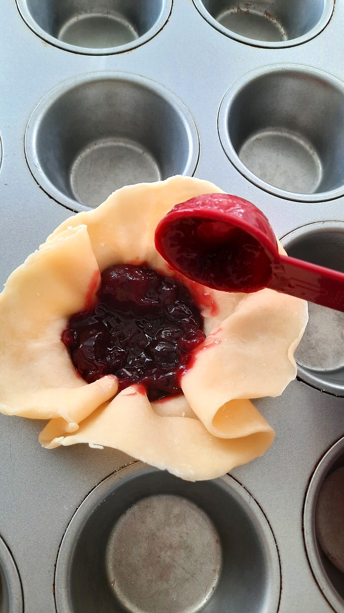a red tablespoon places cherry pie filling into pic crust in muffin tin