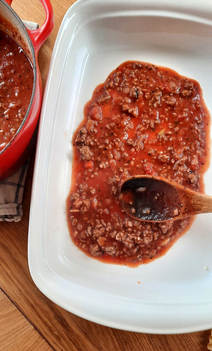 a wooden spoon spreads a layer of tomato meat sauce in the bottom of a 9x13 baking dish