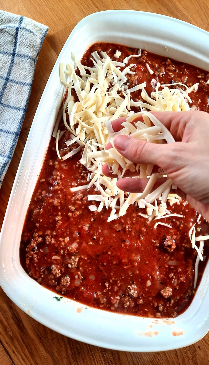 a hand sprinkles grated mozzarella cheese onto uncooked lasagna