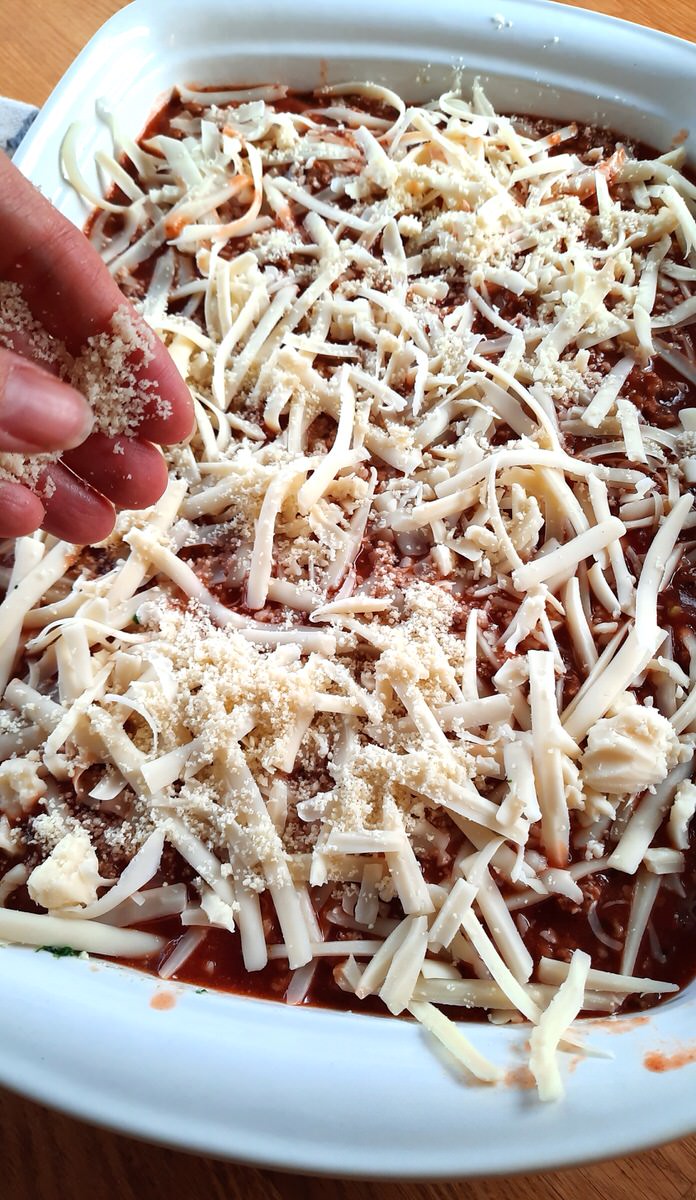 a hand sprinkles parmesan cheese all over the top of homemade lasagna