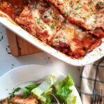 a slice of lasagna on a plate with some salad greens beside the entire dish of homemade lasagna