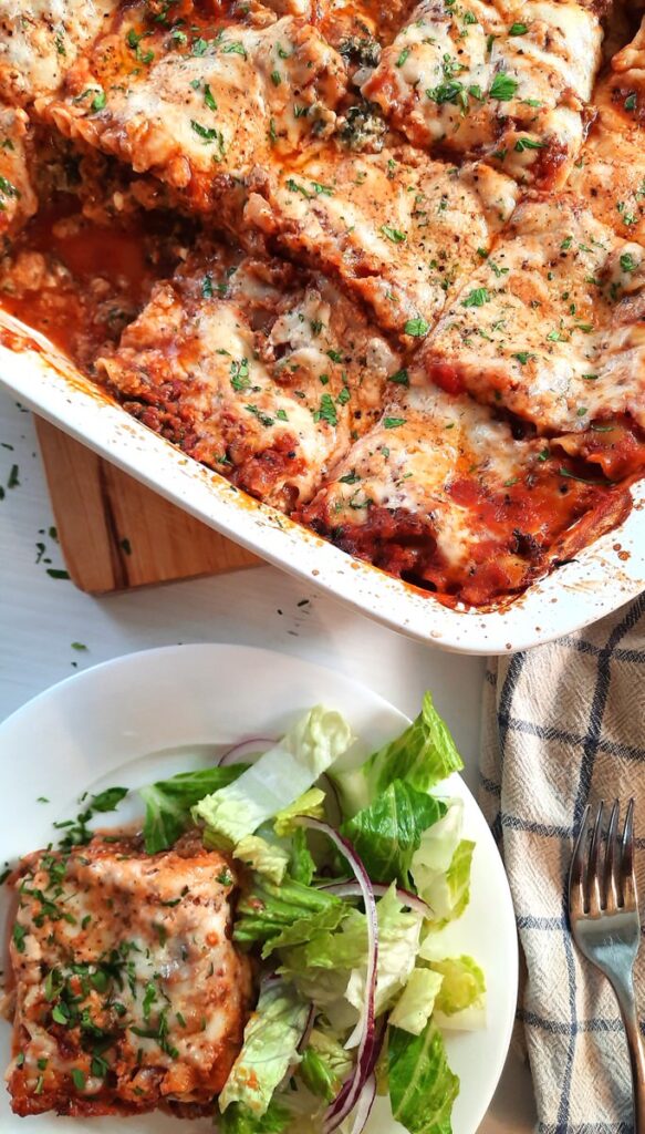 a slice of lasagna on a plate with some salad greens beside the entire dish of homemade lasagna