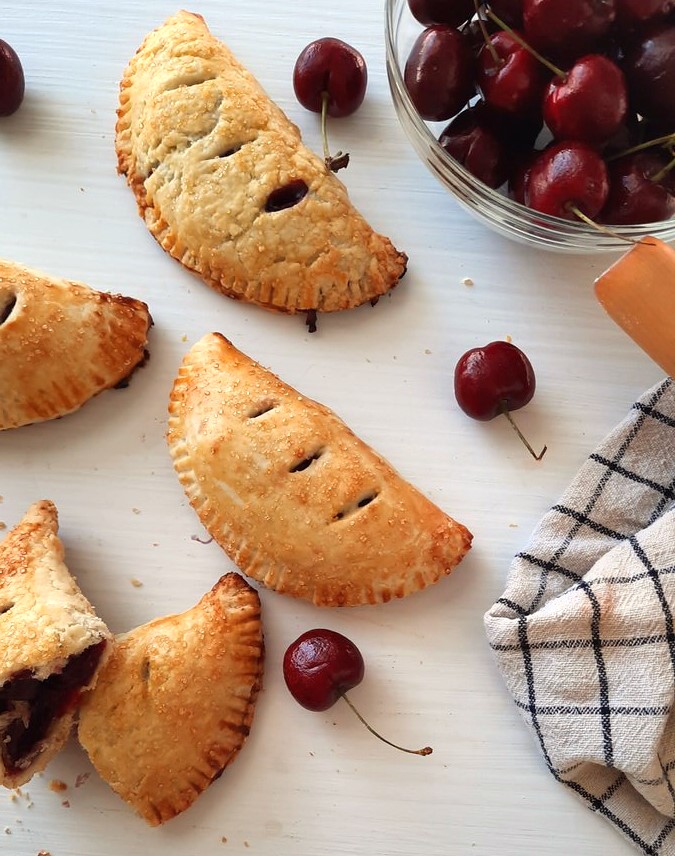 cherry hand pies scattered on a white counter beside a bowl of cherries