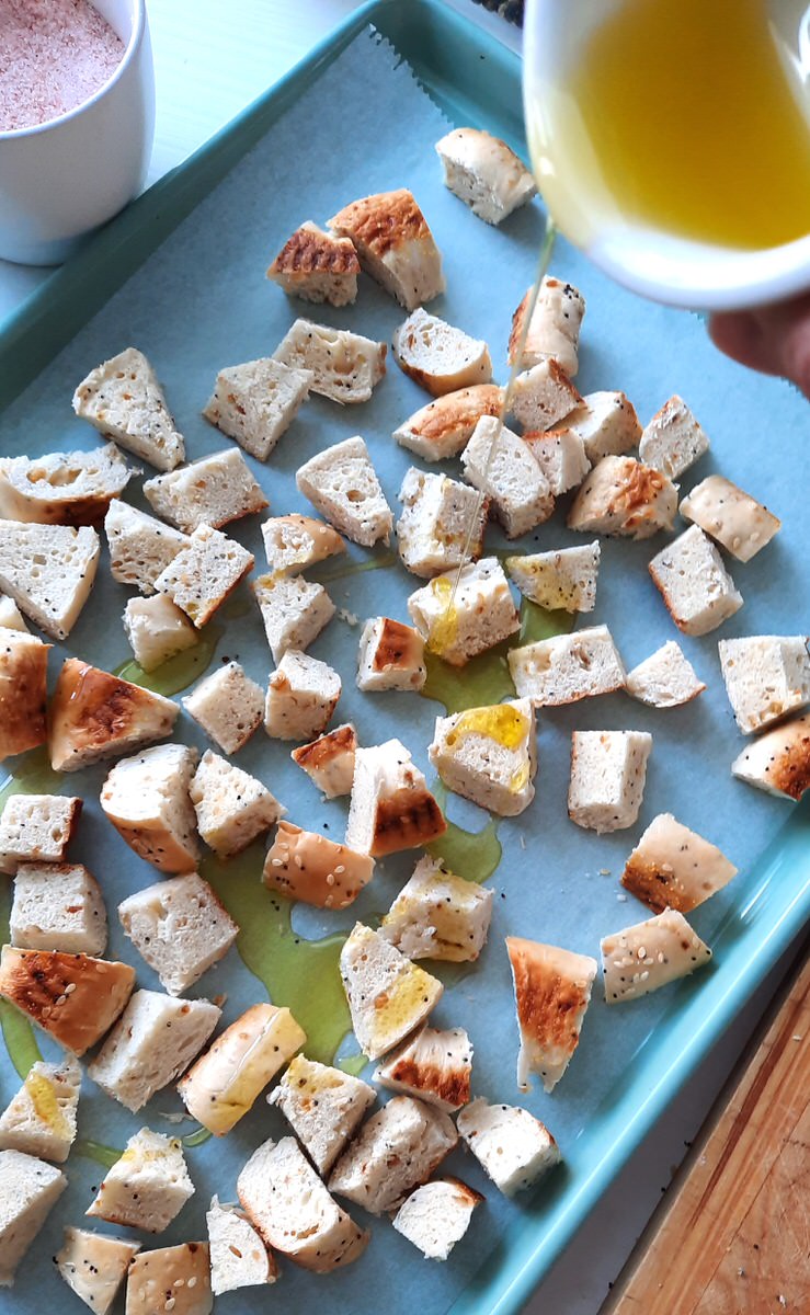 drizzling olive oil onto chopped up bagel croutons on a green baking sheet