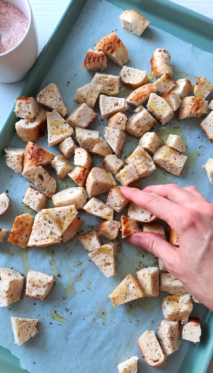 a hand tosses uncooked croutons on a baking sheet